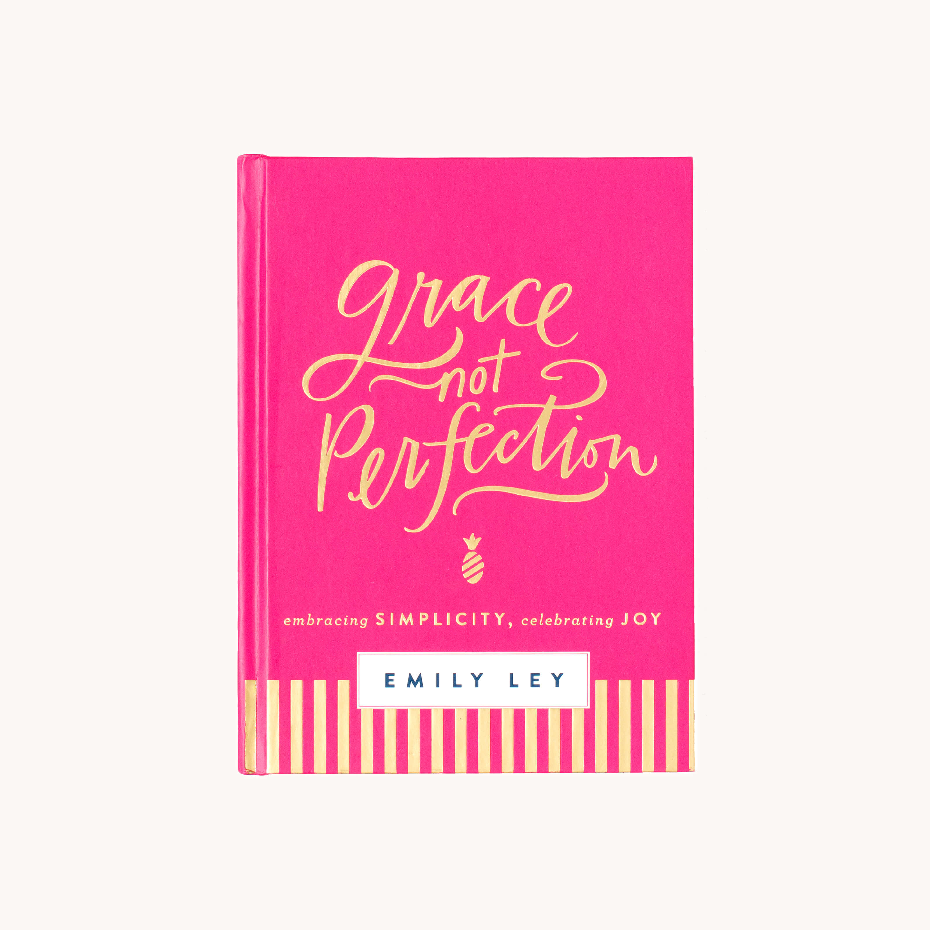 The bright pink book cover, Grace Not Perfection by Emily Ley, features elegant gold script and the encouraging words embracing simplicity, celebrating joy on a white rectangle. Gold stripes at the bottom capture a fulfilling life visual. Its a product of the Simplified brand.