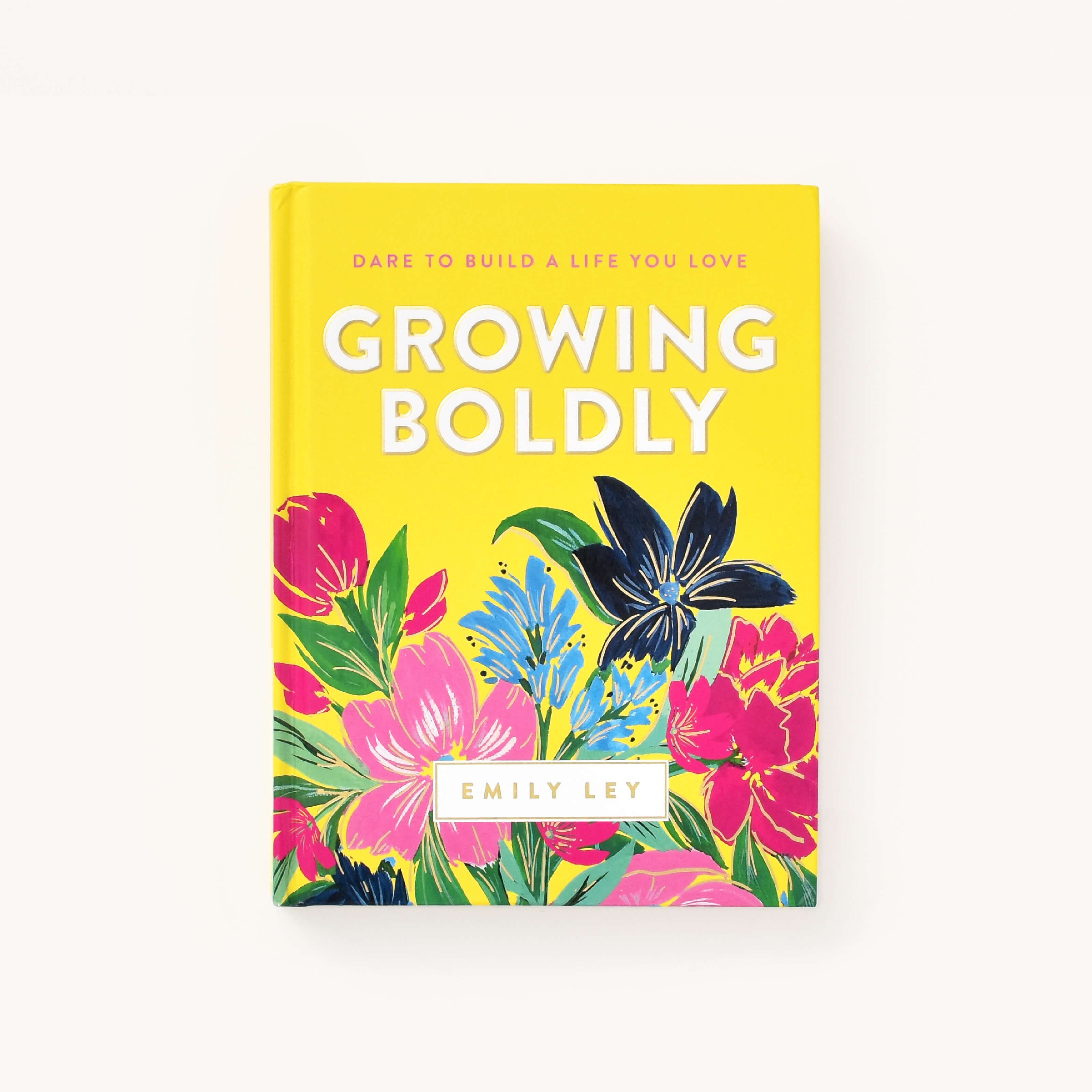 The Simplified book Growing Boldly by Emily Ley features a vibrant yellow cover with illustrations of pink, blue, and black flowers with green leaves. Its tagline, Dare to build a life you love, inspires courage and resilience.