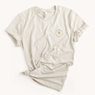 The Grace Not Perfection Tee by Simplified features rolled-up sleeves, a bottom knot, and subtly faded text on the upper left. Its classic beige design laid flat against a white background embodies casual elegance, perfect for any wardrobe.