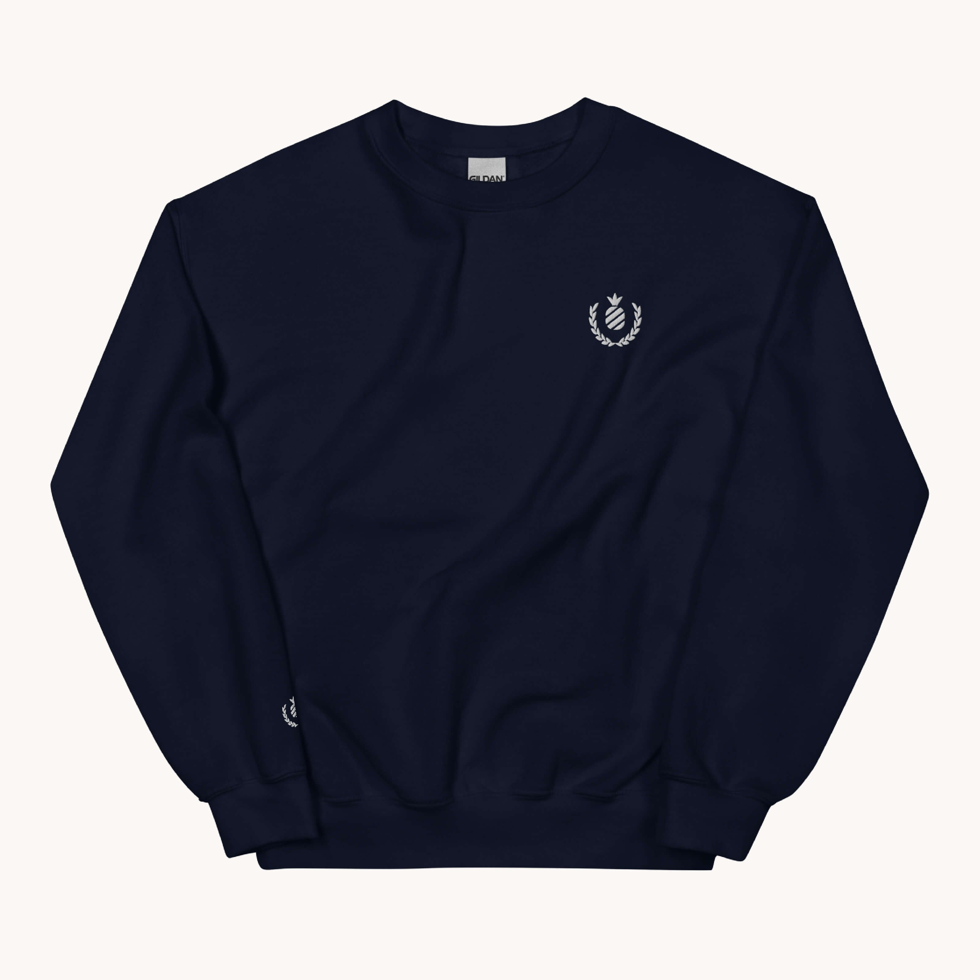 The Pineapple Crest Crewneck by Simplified is a navy blue sweatshirt with a small white laurel wreath and crown emblem on the left chest. Made from soft materials, it features a crew neck, long sleeves, and ribbed cuffs and hem as part of our classic fits apparel collection against a light gray background.