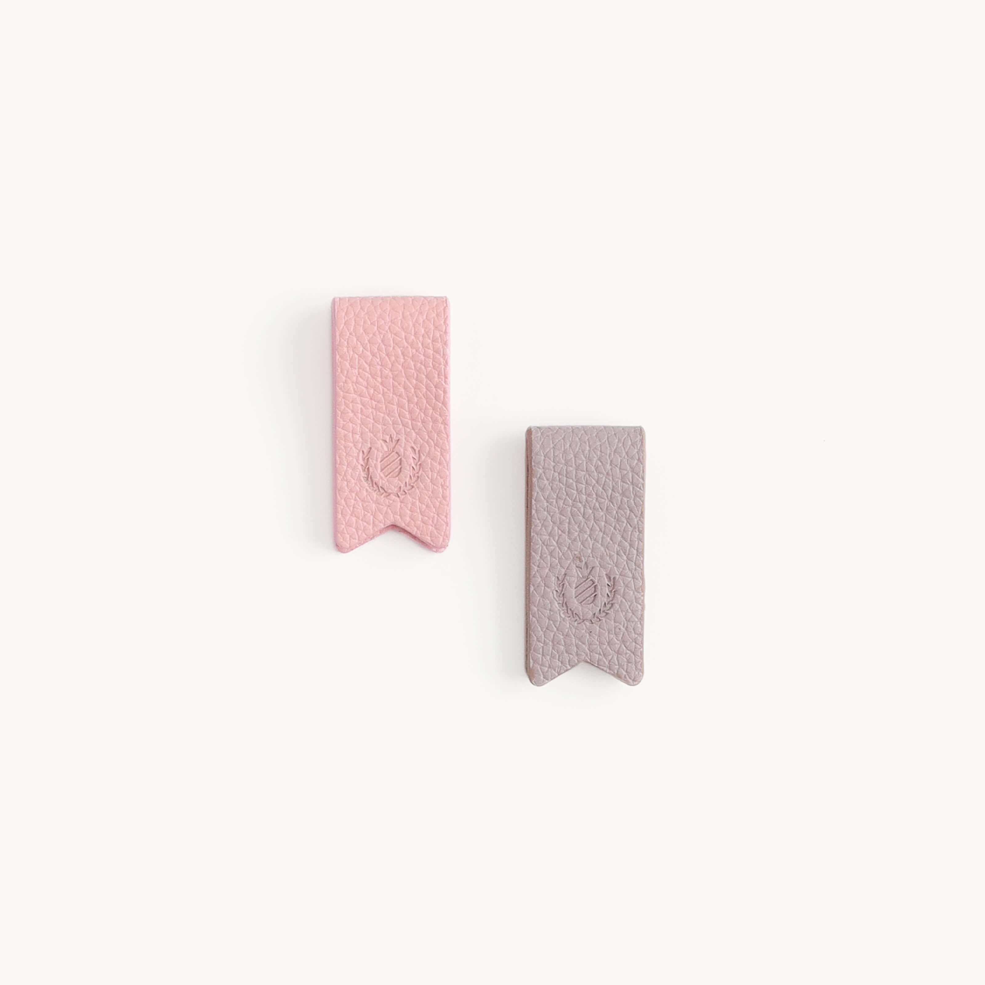 Two Simplified textured leather bookmarks, one blush and one lilac, rest elegantly on a light background. Each features a pointed end and an embossed emblem, reminiscent of their sleek Cord Organizers that bring calm to cable clutter.