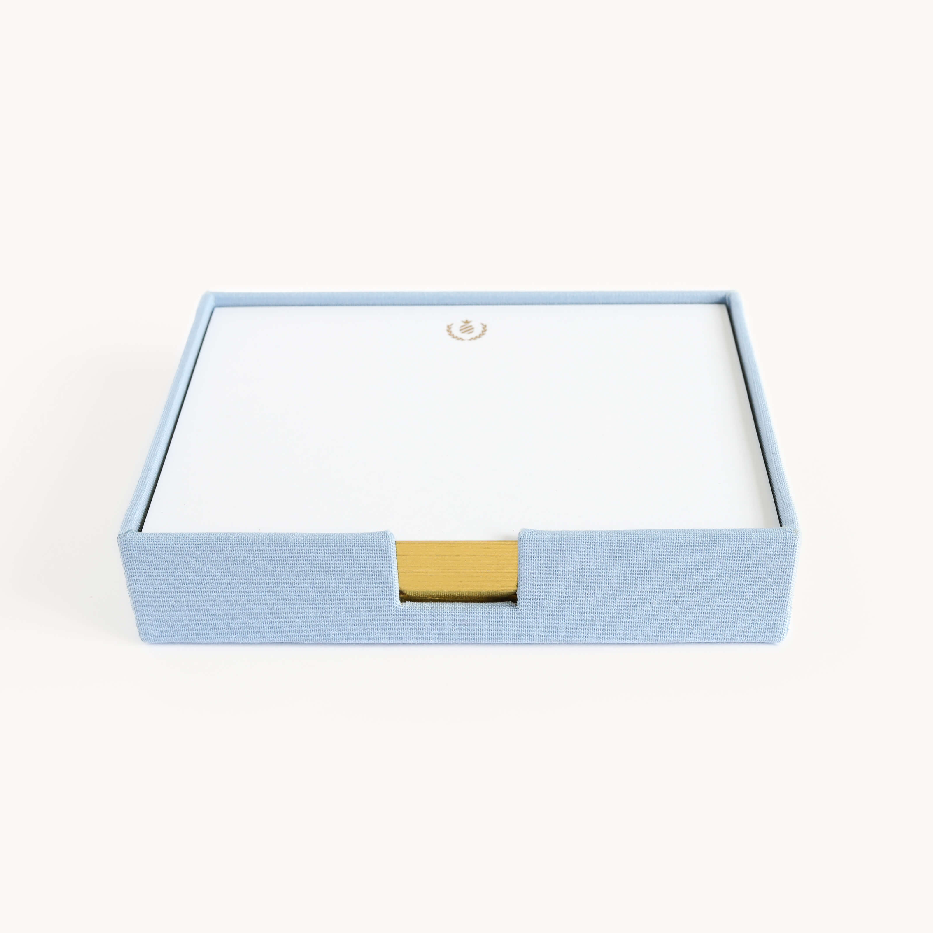 The Desk Jotter - Carolina Linen by Simplified is a square blue linen box with a gold tab, containing gold-gilded notecards featuring a gold wreath emblem. The box is open, showcasing the top card of this refined set.