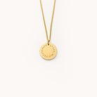 Simplifieds Grace Not Perfection gold necklace features a round engraved pendant on a thin chain, exuding elegance and simplicity against a light background.