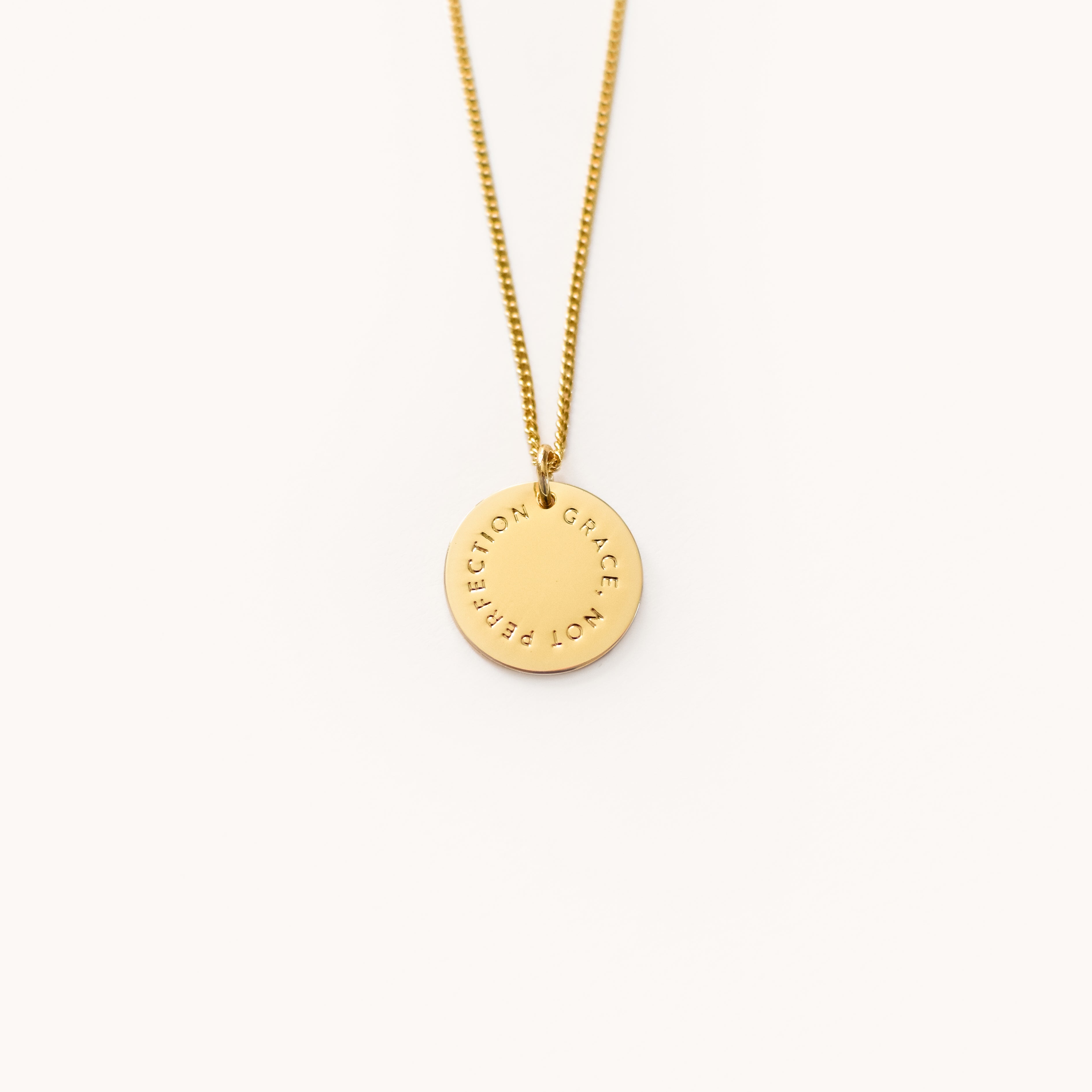 Simplifieds Grace Not Perfection gold necklace features a round engraved pendant on a thin chain, exuding elegance and simplicity against a light background.