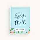 The image shows the hardcover book When Less Becomes More by Emily Ley, from the Simplified brand. The minimalist cover is light blue with elegant script and decorated with pink flowers and greenery.