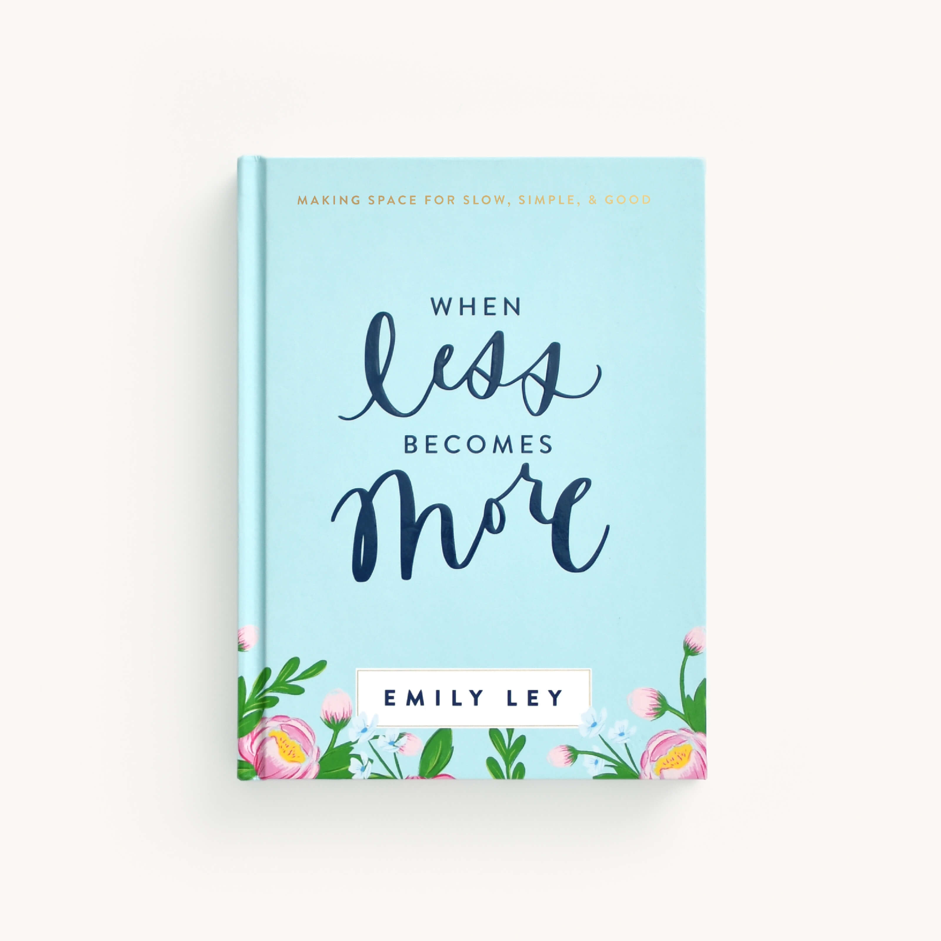 The image shows the hardcover book When Less Becomes More by Emily Ley, from the Simplified brand. The minimalist cover is light blue with elegant script and decorated with pink flowers and greenery.