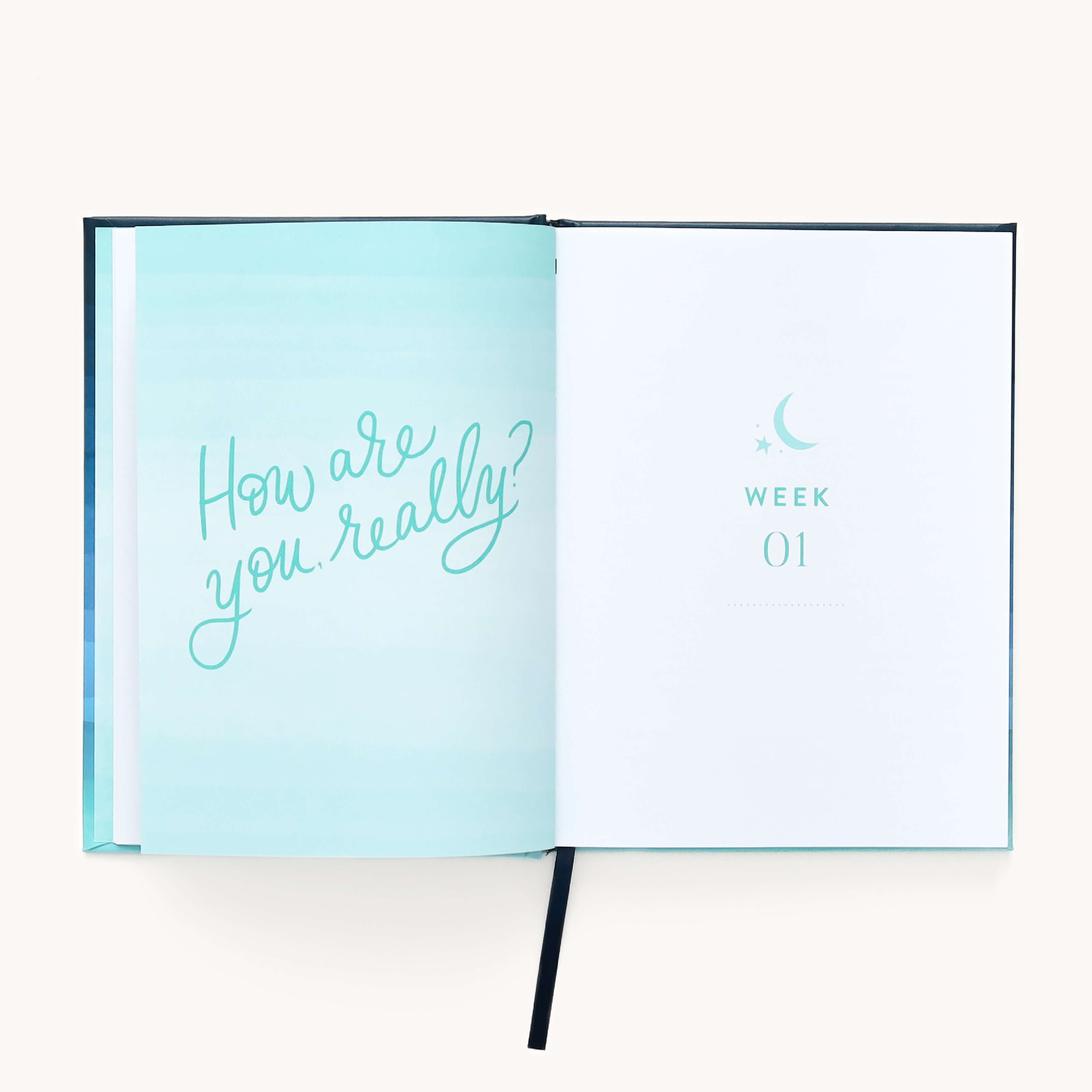 The Near in the Night journal by Emily Ley for Simplified features an open spread; the left page in teal asks, How are you, really? for reassurance. The right page shows WEEK 01 with a crescent moon and stars, creating calm. An elegant black ribbon bookmark lies at the bottom.