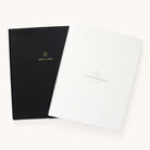 Two sleek Simplified planners with leatherette covers are displayed. The black one features gold 2024 to 2025 text, while the white one reads Dapperdesk by Emily Ley. Crafted from high-quality paper, the planners slightly overlap on a plain background.