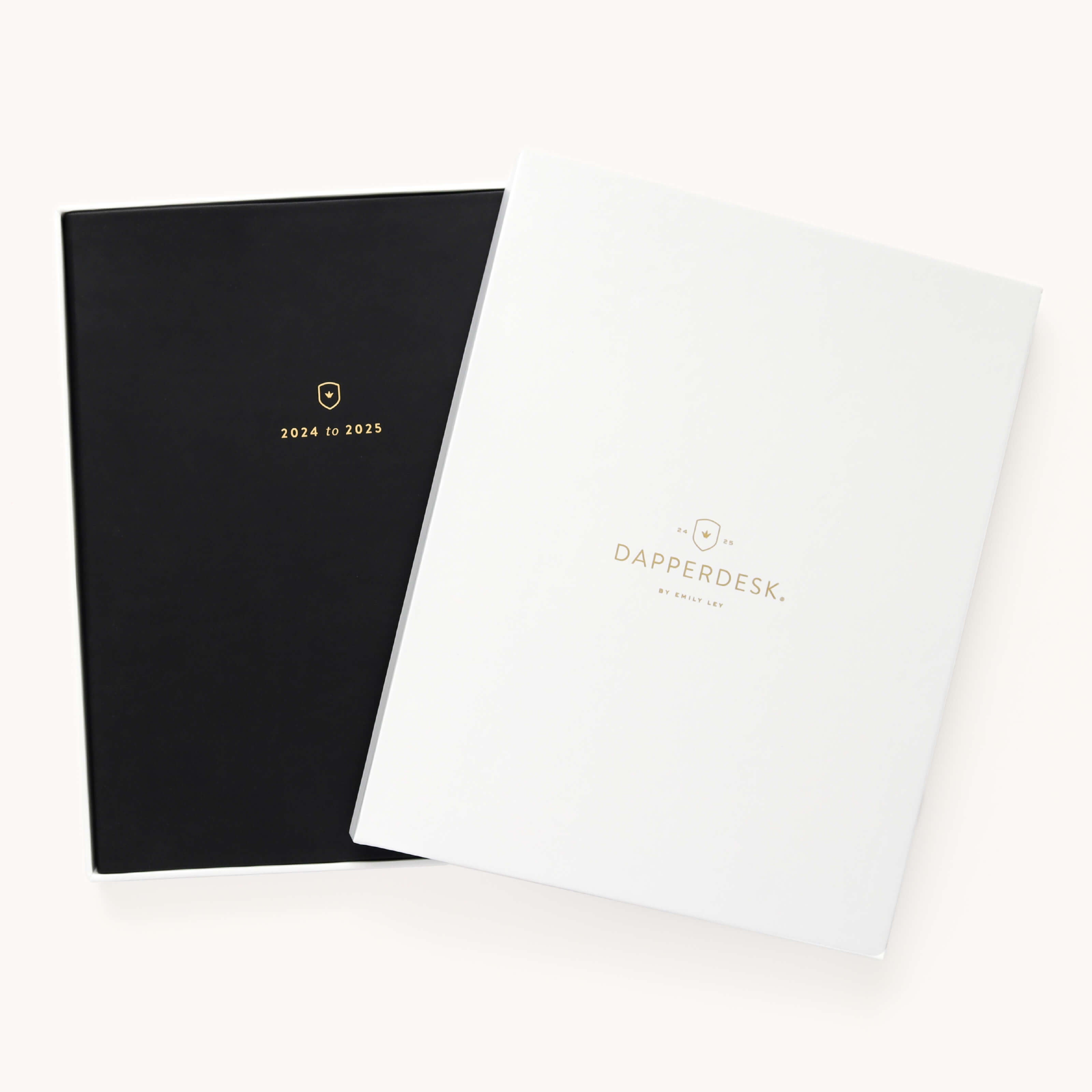 Two sleek Simplified planners with leatherette covers are displayed. The black one features gold 2024 to 2025 text, while the white one reads Dapperdesk by Emily Ley. Crafted from high-quality paper, the planners slightly overlap on a plain background.