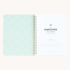 Simplifieds Workbook - Project Management is an open spiral-bound notebook with a patterned teal and white cover featuring geometric shapes. It includes lined pages and monthly spreads for seamless planning and organization.