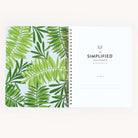 An open spiral-bound Workbook - Travel Planning from Simplified lies flat, featuring green tropical leaves on a light blue background on the left page. The right page is titled The Simplified Travel Workbook, offering space for your name—ideal for organizing itineraries and travel plans effortlessly.