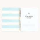 An open spiral-bound wellness workbook by Simplified is shown. The left page displays blue and white stripes, while the right page invites goal tracking with a Belongs To section beneath the title Workbook - Wellness.