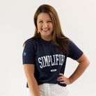 A woman with long brown hair wears a Simplified Collegiate Tee in navy blue and classic fit white pants. She stands against a plain white background, epitomizing timeless style from the latest apparel collection by Simplified.