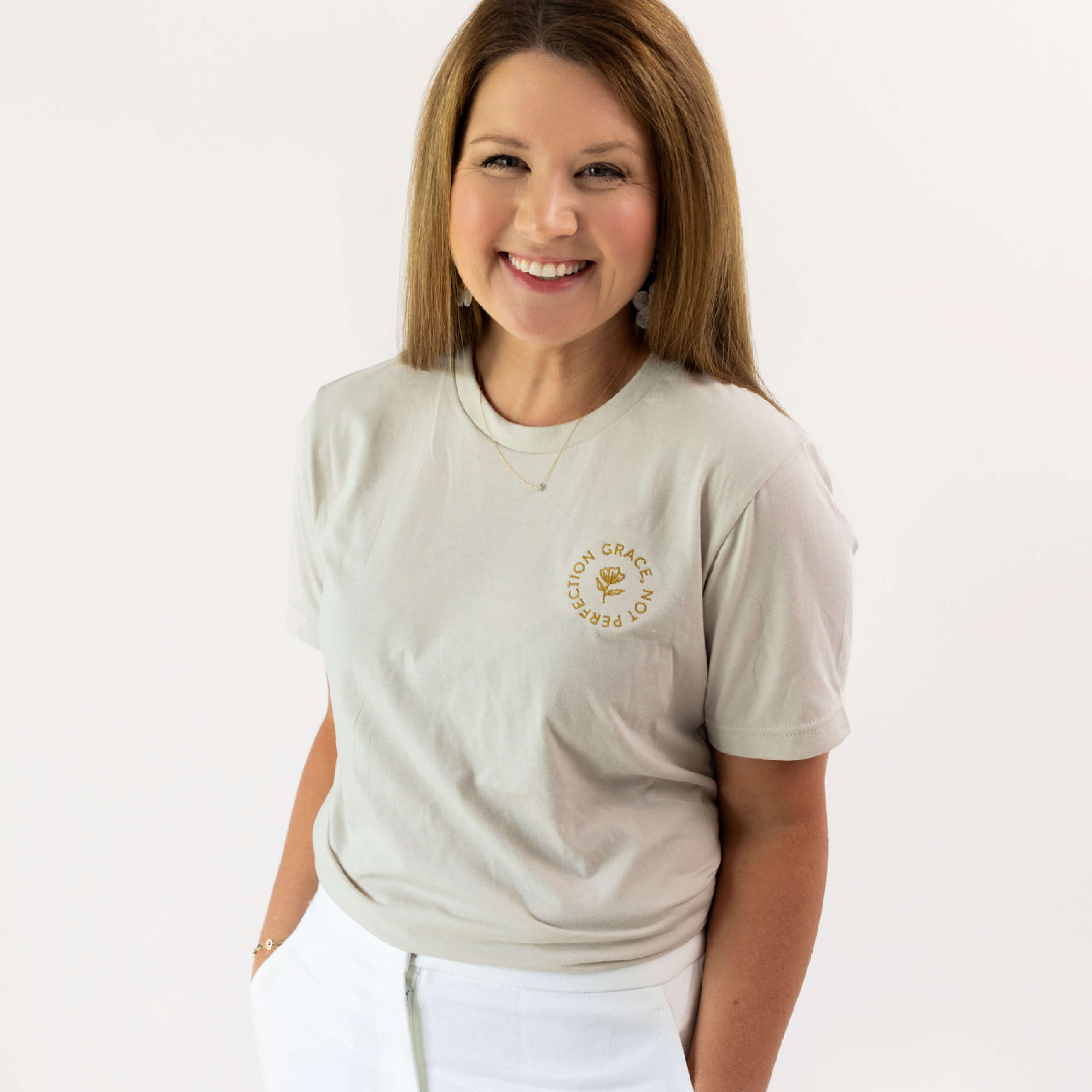 A person with long brown hair smiles at the camera, showcasing an apparel collection. They wear a Grace Not Perfection Tee by Simplified in light gray with text on the chest and classic fit white pants against a plain white background.