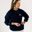 A person with long brown hair wears a Simplified Pineapple Crest Crewneck, navy blue from the classic fits collection. They accessorize with gold floral earrings, a light blue headband, and a bracelet. Against a plain white background, they smile with one hand on their hip.