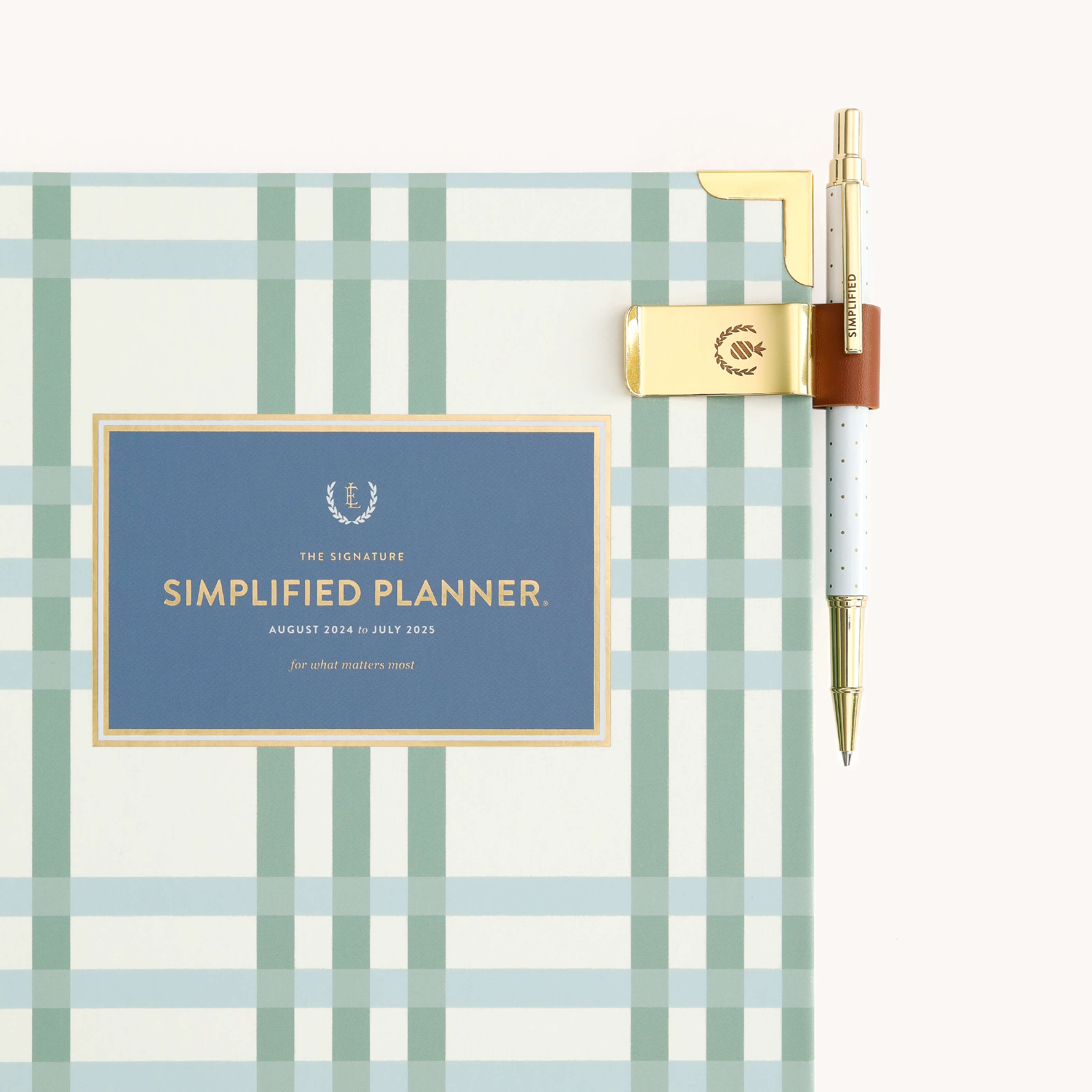 A plaid-patterned planner titled Simplified Planner, covering August 2024 to July 2025, includes a saddle pen clip securing an elegant white pen with a gold tip at the top right corner.