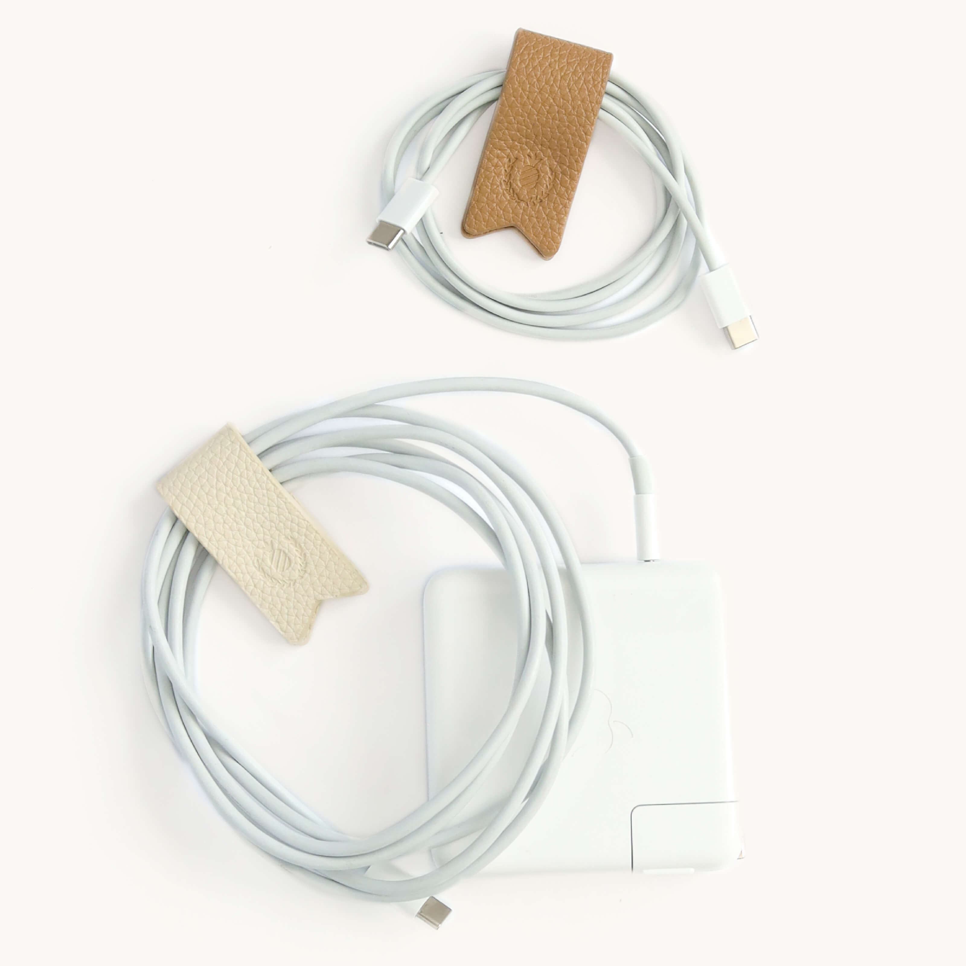 Two coiled white USB-C cables are secured by Simplifieds Cord Organizers in Ivory & Fawn. The smaller tan and the larger beige cables, attached to a white charger, rest neatly on a plain white background, eliminating cable clutter with stylish organization.