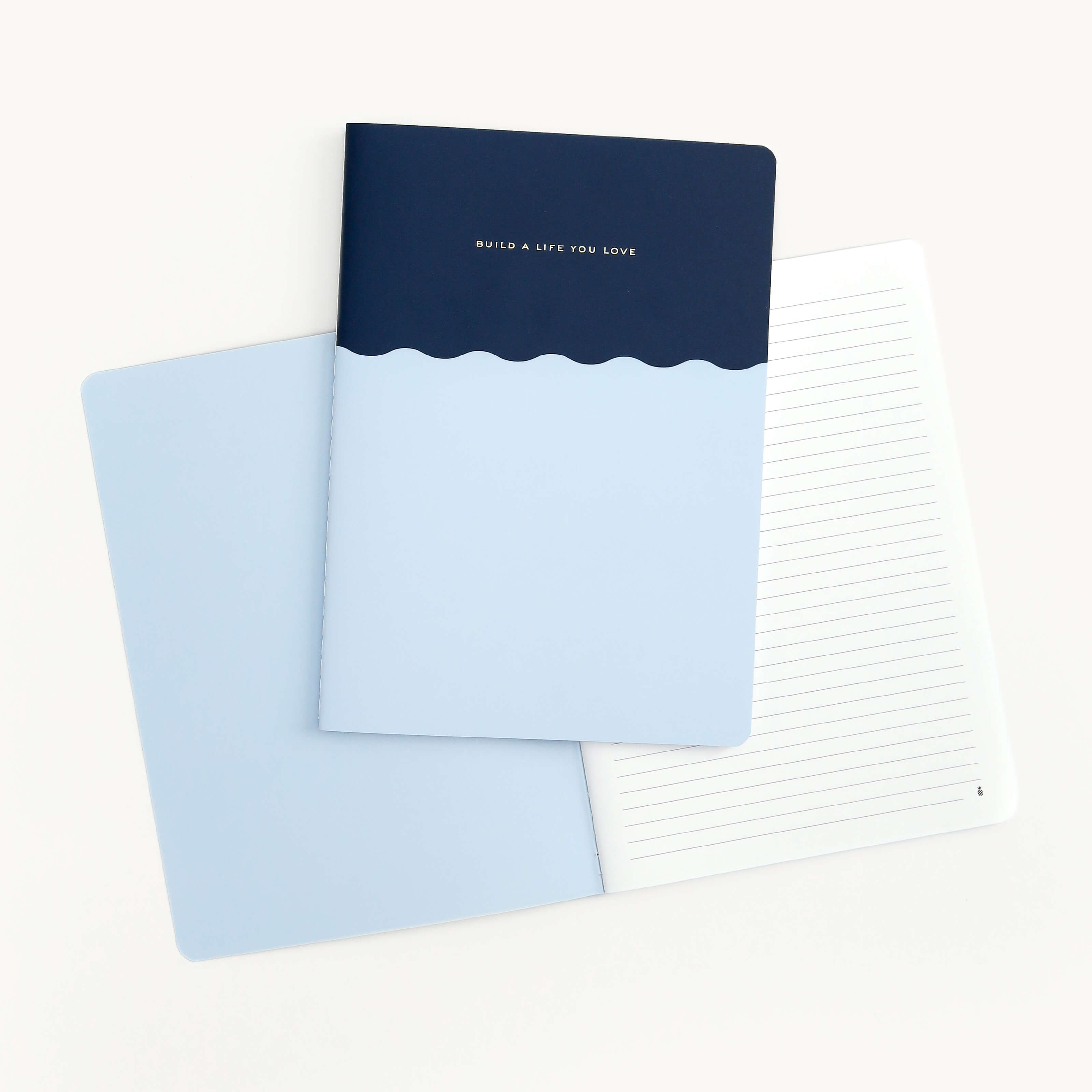 Three mid-size notebooks from Simplified are showcased on a white background: one features a dark blue cover inscribed with BUILD A LIFE YOU LOVE and a light blue wave motif, while the others are open to display their blank and lined pages, all elegantly saddle-stitched.