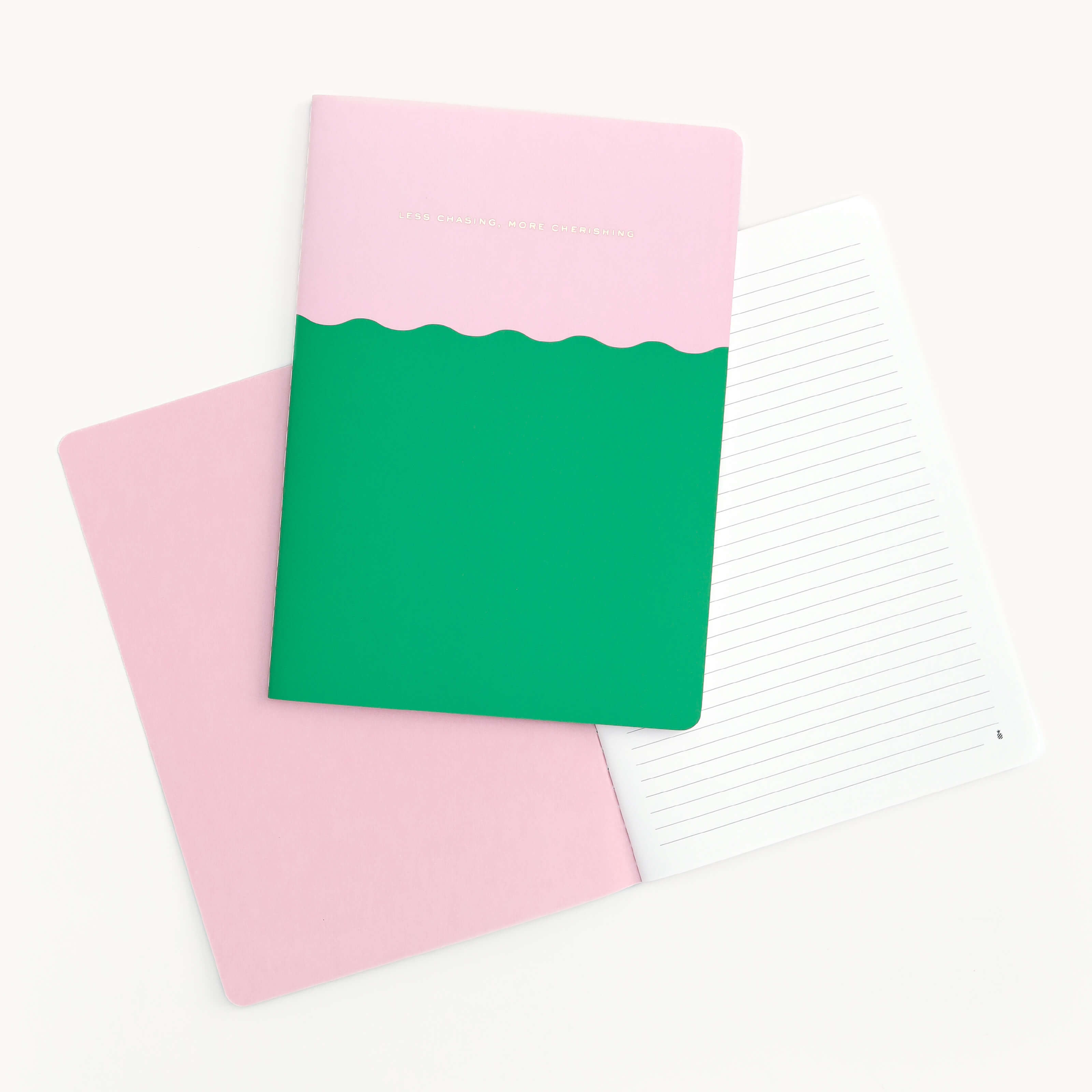 Two pastel notebooks are shown. On the left, the closed mid-size Less Chasing More Cherishing notebook by Simplified has a pink top, green bottom, wavy border, and saddle-stitched binding. Its elegant design complements the open pink one with lined pages on a cream background.