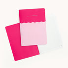 Two Simplified Mid-Size Notebooks are shown: one closed with Grace, Not Perfection on the cover and a wavy design dividing two pink shades, featuring saddle-stitched binding; the other is open to lined pages.