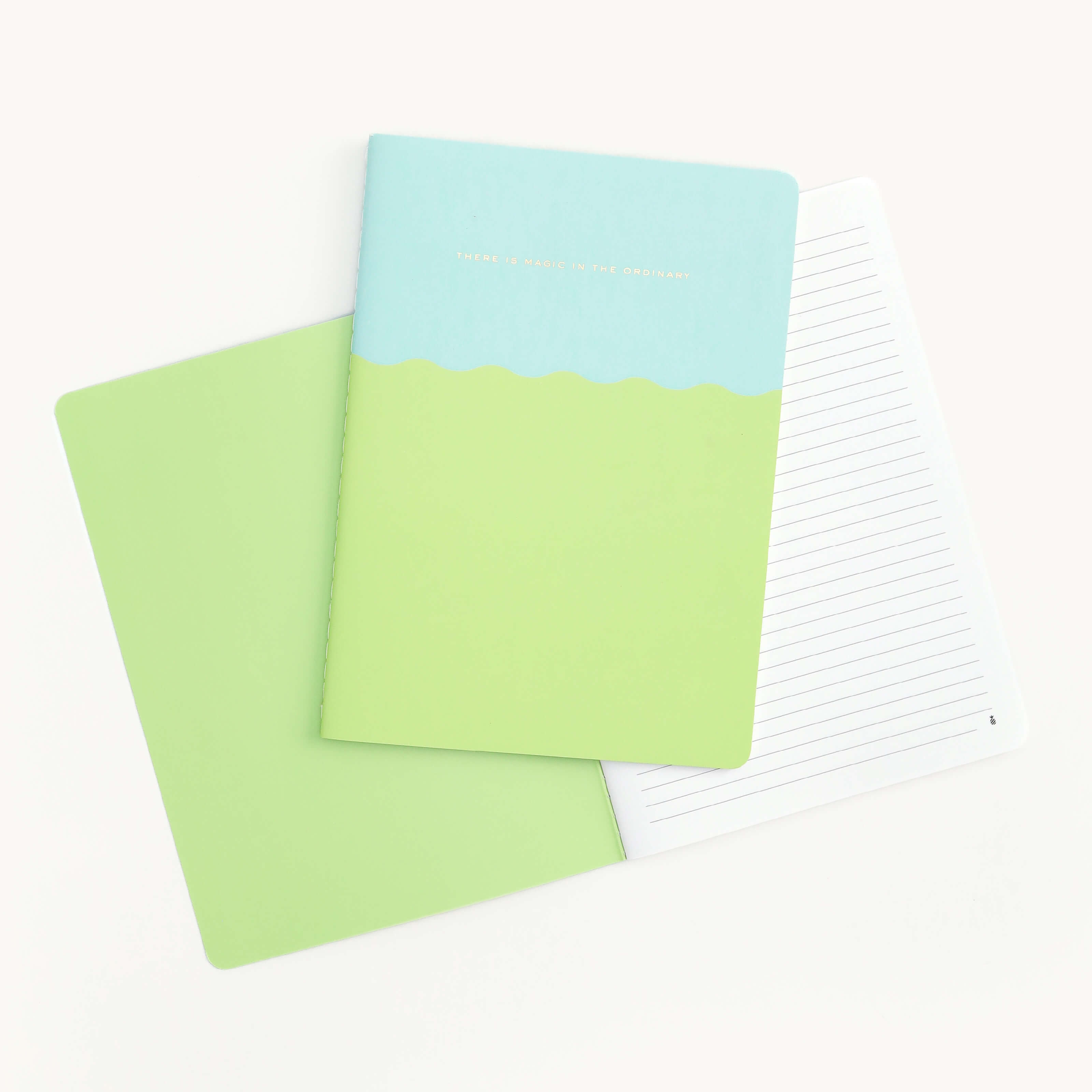 Two Mid-Size Notebooks by Simplified, Magic In The Ordinary, are displayed with colorful covers: one in a blue-green wavy design and the other solid green. They feature saddle-stitched binding, and opened notebooks reveal lined pages.