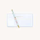 A floral pen diagonally atop Simplifieds Notepad - Habit Tracker, ideal for reminders. The light blue tracker has grid sections for days and tasks, with Habit Tracker elegantly in gold at the top.