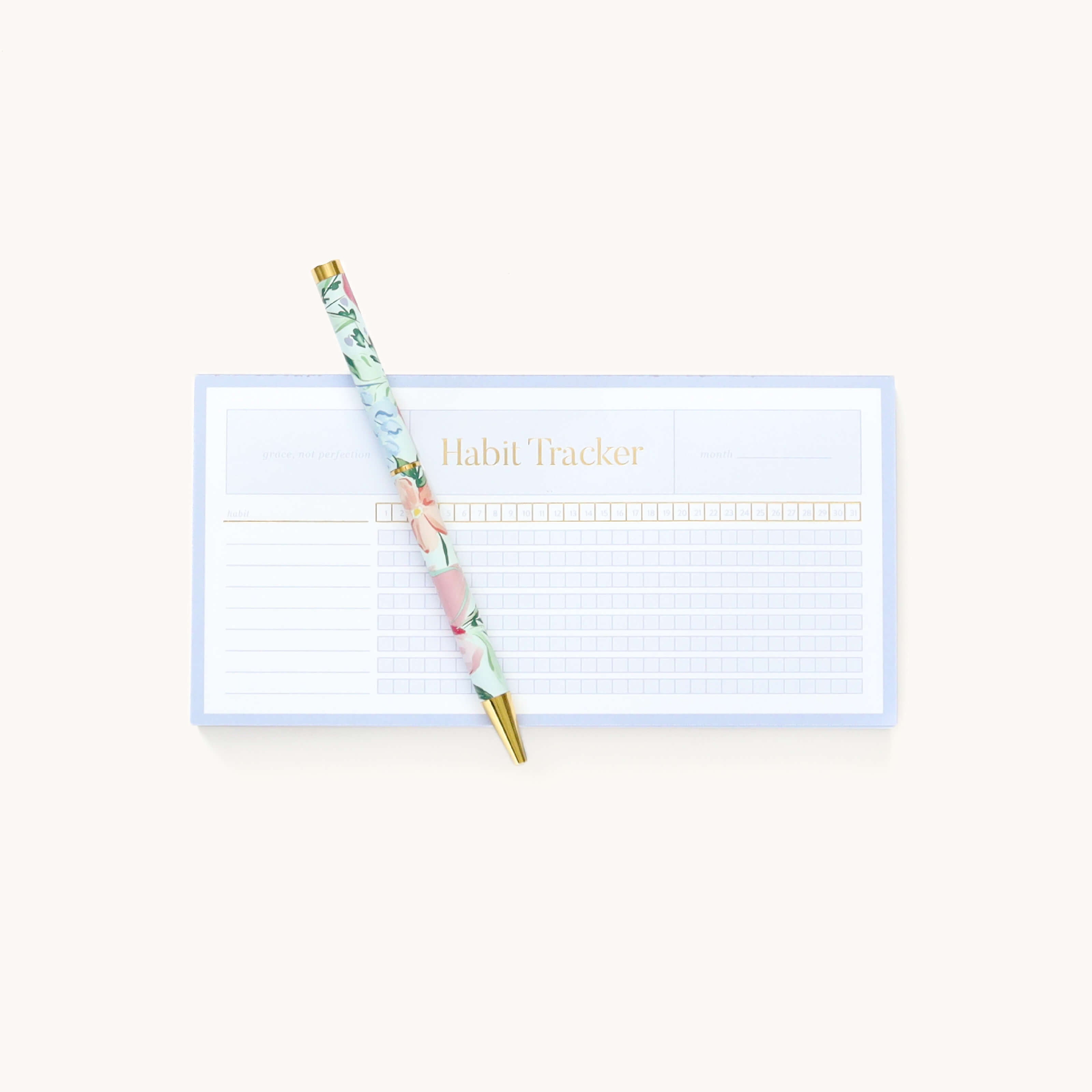 A floral pen diagonally atop Simplifieds Notepad - Habit Tracker, ideal for reminders. The light blue tracker has grid sections for days and tasks, with Habit Tracker elegantly in gold at the top.