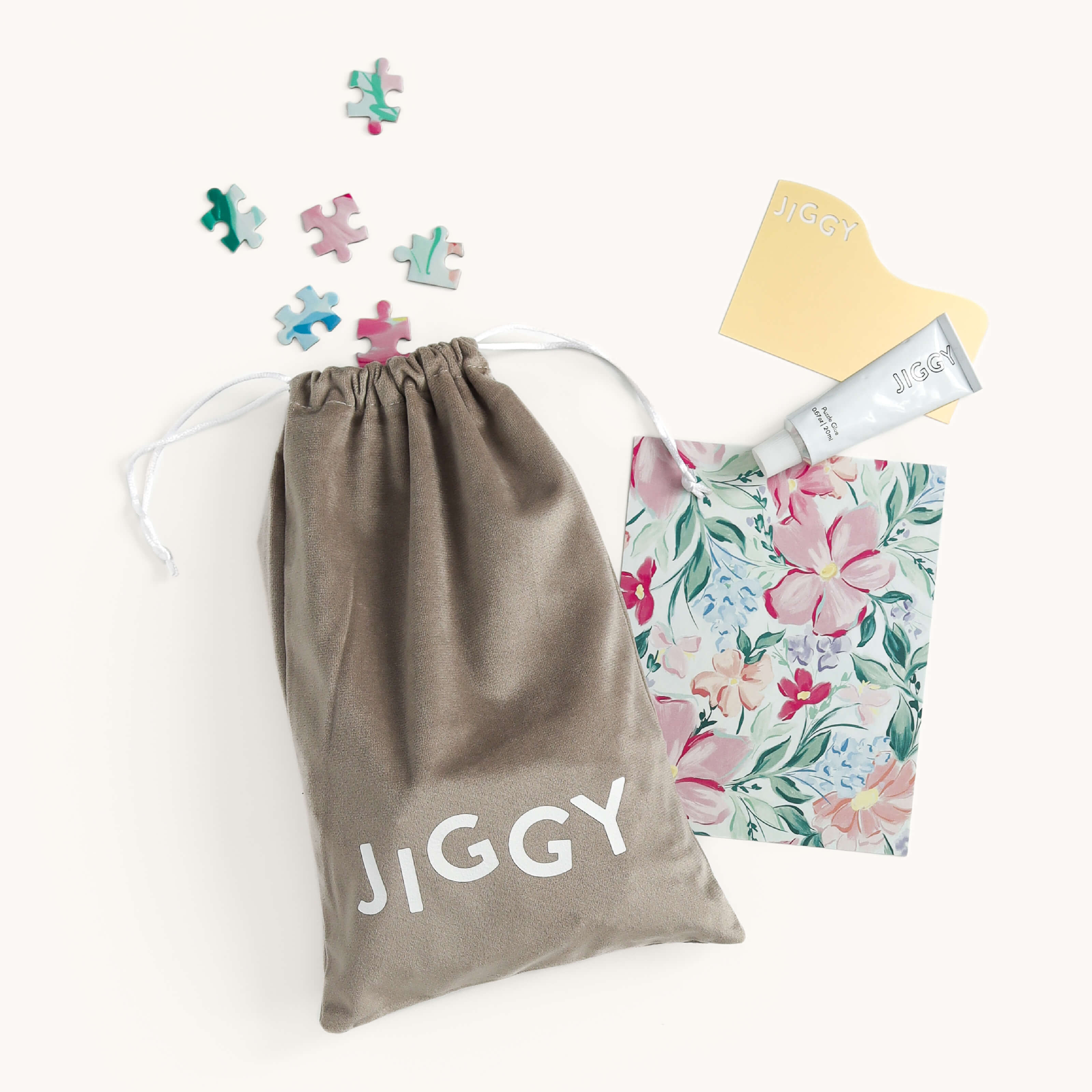 A gray drawstring bag with JIGGY, scattered puzzle pieces, and a Blush Magnolia Jiggy Puzzle box (by Simplified) create a charming scene. A branded tube and a JIGGY card enhance the watercolor floral theme against its light backdrop.