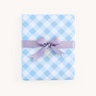 A present wrapped in Simplifieds vibrant Carolina Gingham sheets, accented with a purple ribbon bow, sits elegantly against a plain white backdrop.