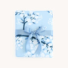 A Simplified gift wrapped in Cherry Blossoms wrapping sheets, featuring light blue floral patterns with white and navy accents. Its adorned with a matching satin blue ribbon and bow, all set against a plain white background.