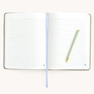 The Leatherette Journal - Fawn by Simplified lies flat with an elegant blue ribbon bookmark centered, a gold and green striped pen diagonally on the right page, against a plain backdrop highlighting subtle gold foil details.