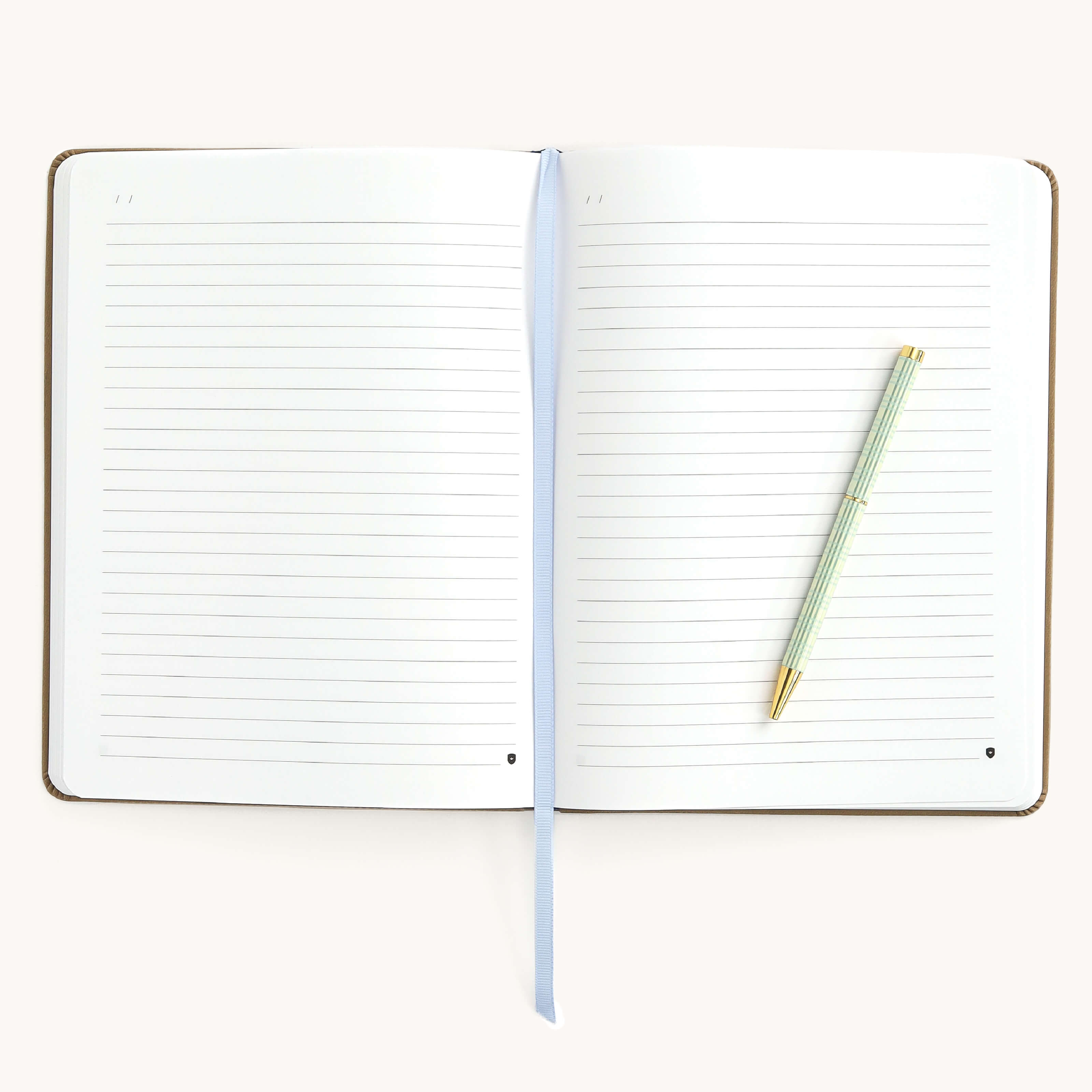 The Leatherette Journal - Fawn by Simplified lies flat with an elegant blue ribbon bookmark centered, a gold and green striped pen diagonally on the right page, against a plain backdrop highlighting subtle gold foil details.