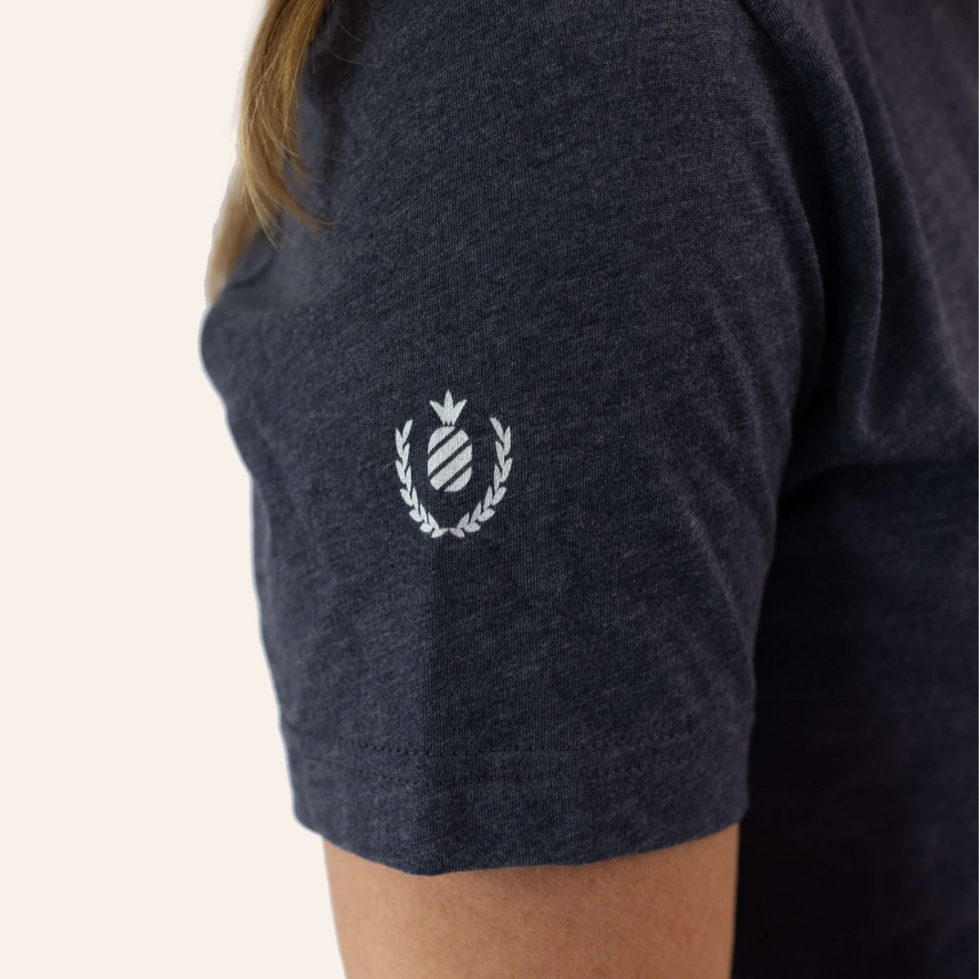 A person wears the Simplified Collegiate Tee in dark gray, featuring a small white emblem on the sleeve. The design includes a circle surrounded by a wreath, highlighting classic fits. A plain light background emphasizes its soft materials.