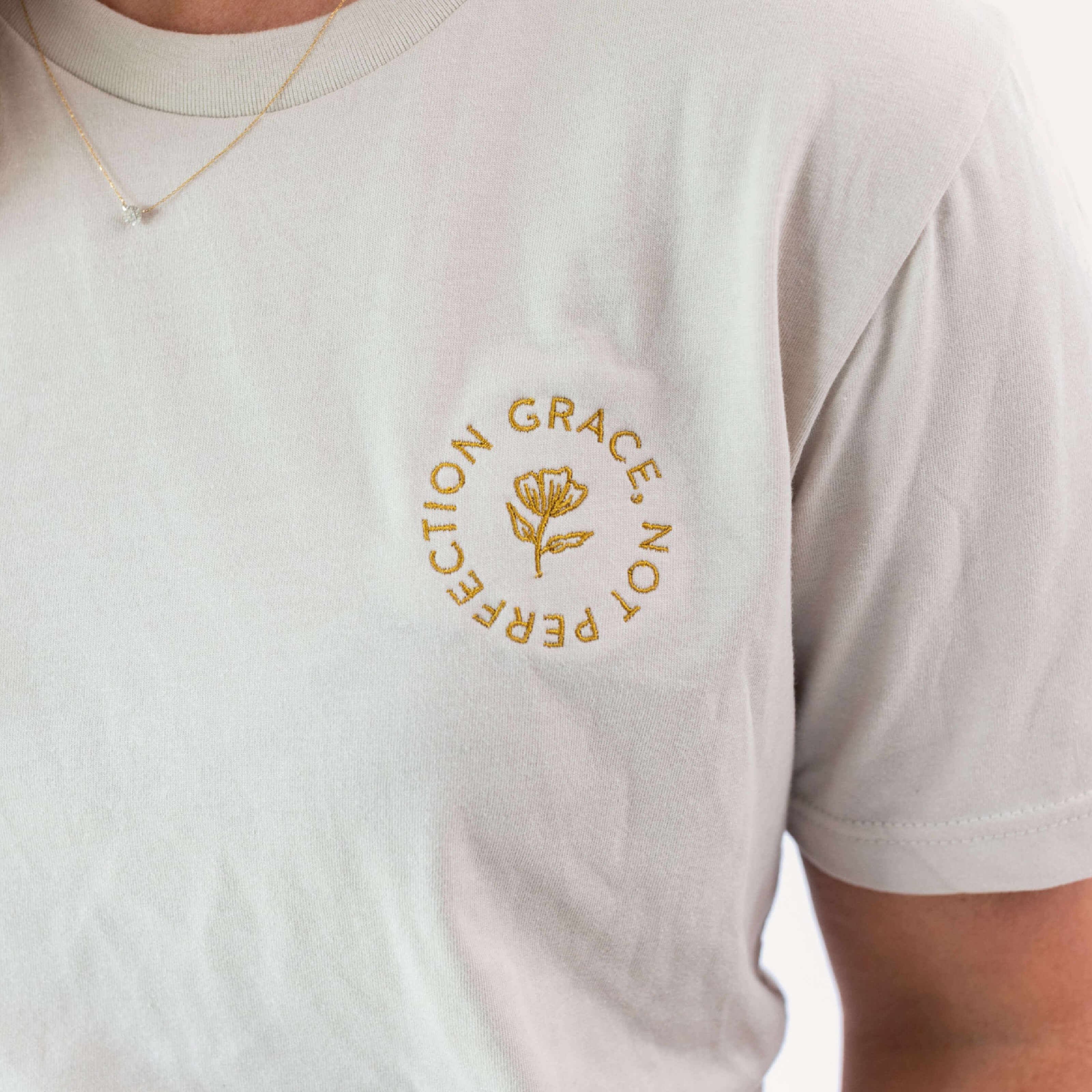 Close-up of a person wearing Simplifieds Grace Not Perfection Tee in light beige, showcasing a gold embroidered flower encircled with GRACE NOT PERFECTION on the chest. A delicate necklace complements this design.