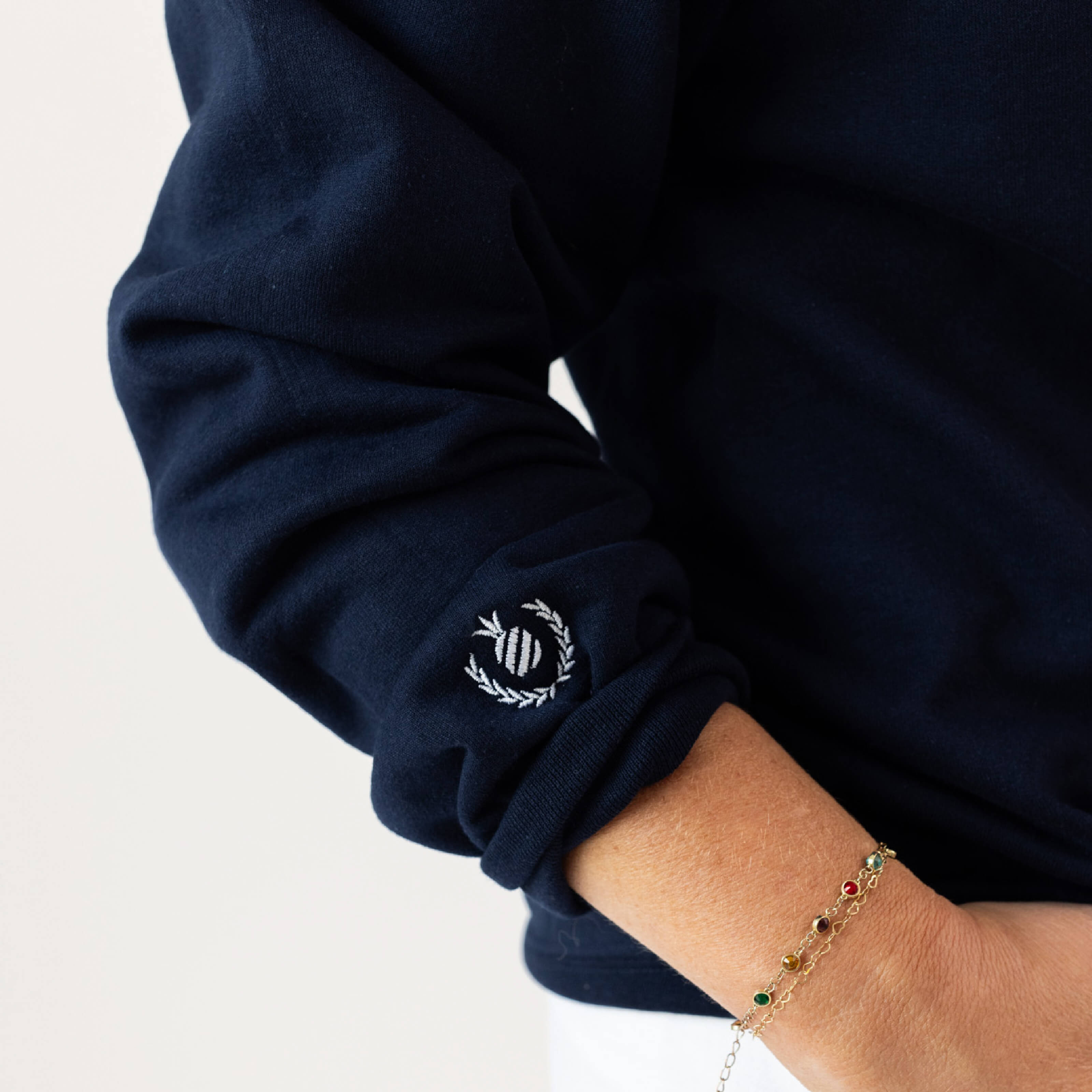 A person is wearing a Pineapple Crest Crewneck by Simplified, featuring navy fabric with an embroidered design near the sleeve. Their hand, resting on their hip, is adorned with a delicate chain bracelet showcasing colorful gemstones against a plain white background.