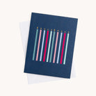 Simplifieds Boxed Card Set features an elegant greeting card with a dark blue cover and nine lit candles in pink, light blue, and red. Perfect for any occasion, it comes partially inside a crisp white envelope.