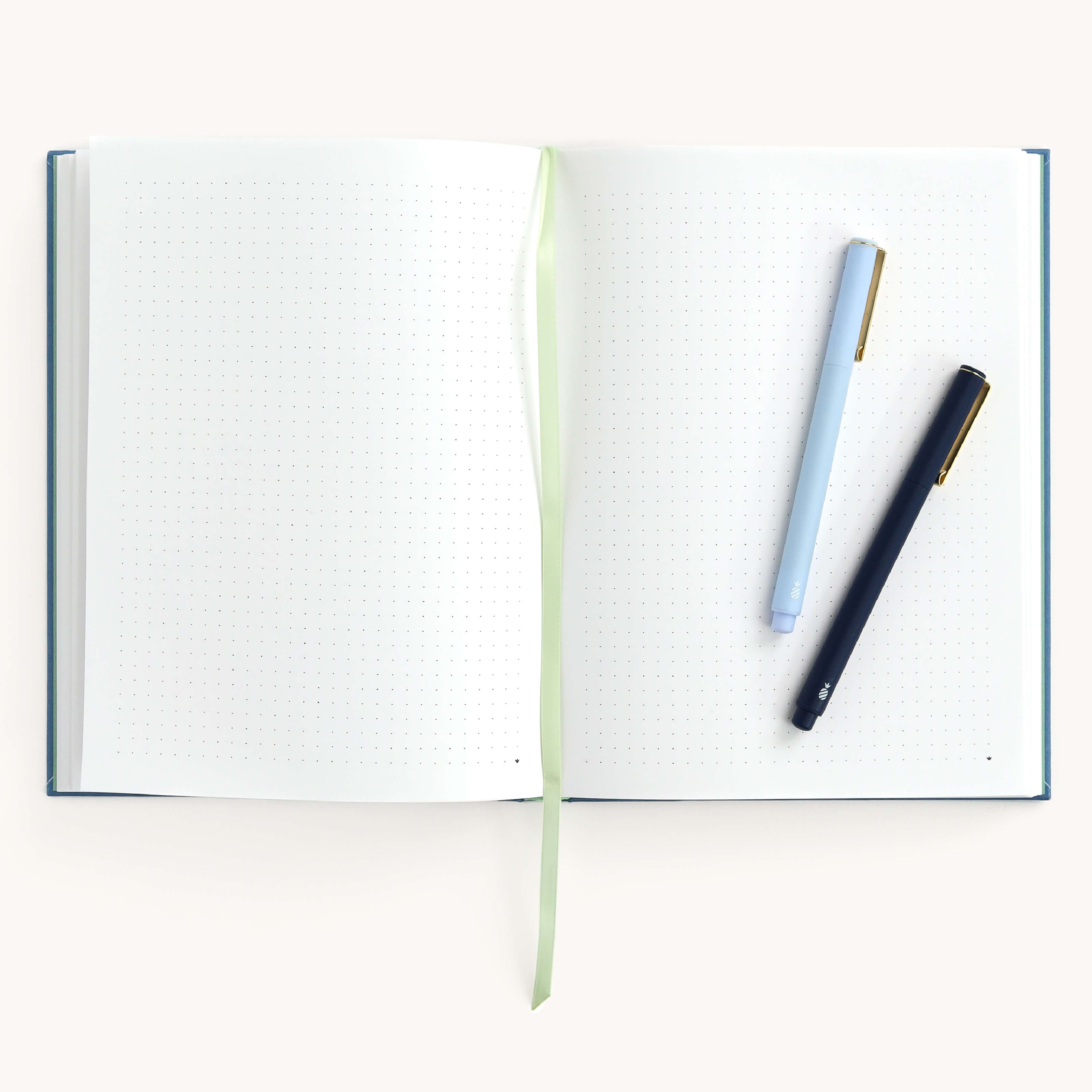 The Simplified Bullet Journal in French Blue features an open linen cover with dot-grid pages that lay flat. A light green ribbon marker is centered, while a light blue and dark blue pen rest diagonally on the right page against a white background, ideal for bullet journaling.