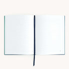 The Linen Journal - Navy by Simplified lies open with lined pages on a white surface, featuring a dark ribbon bookmark between pages. Its simple design with a visible spine invites reflection and creativity.