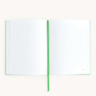 The Linen Journal - Sage by Simplified, featuring lined pages and a green bookmark ribbon at its center, invites creativity and reflection against a plain white backdrop.