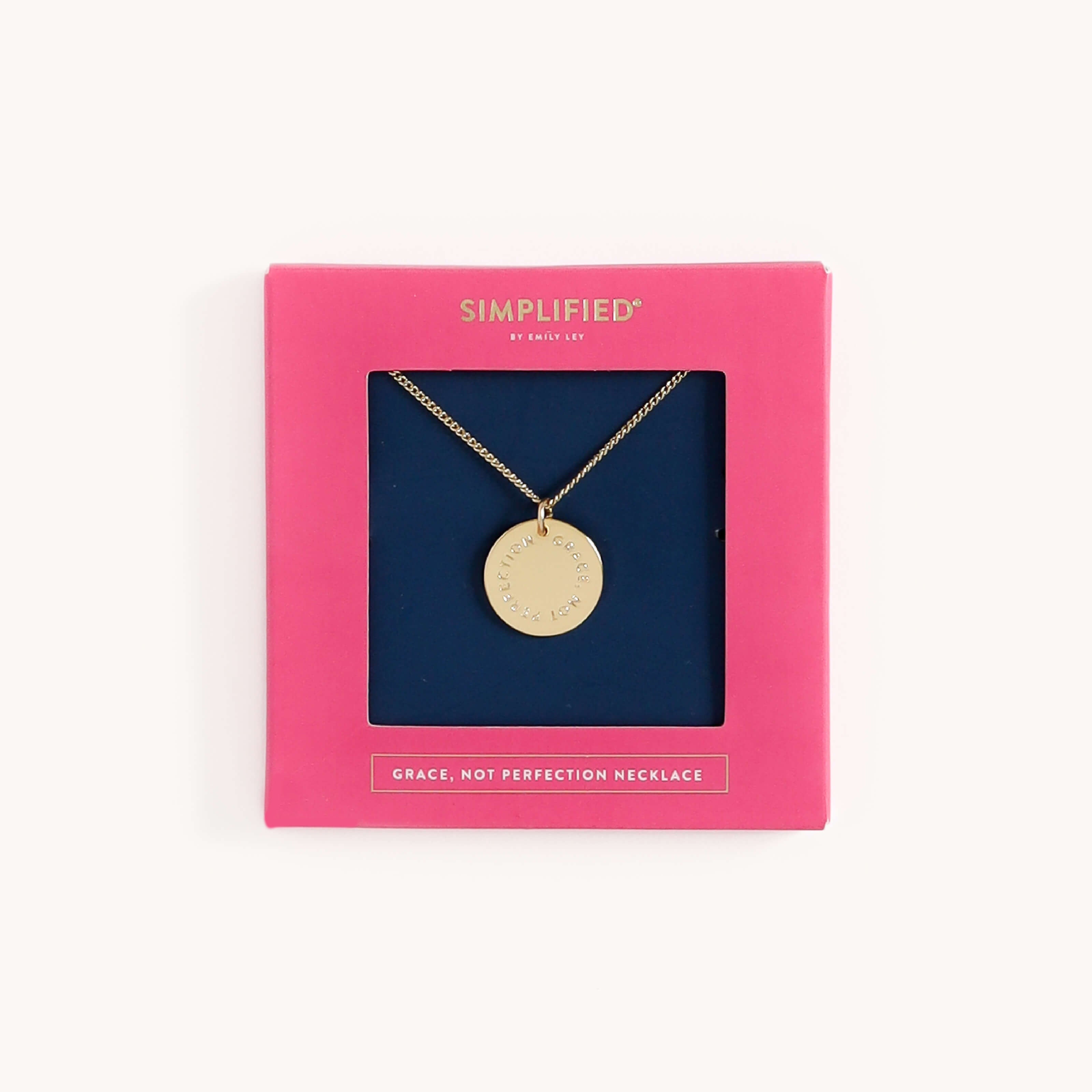 The Necklace - Grace Not Perfection by Simplified features a gold piece with a round pendant inscribed GRACE, NOT PERFECTION, exuding elegance. It’s presented in a pink SIMPLIFIED box that captures simplicity, bearing the inscription GRACE, NOT PERFECTION NECKLACE at the bottom.