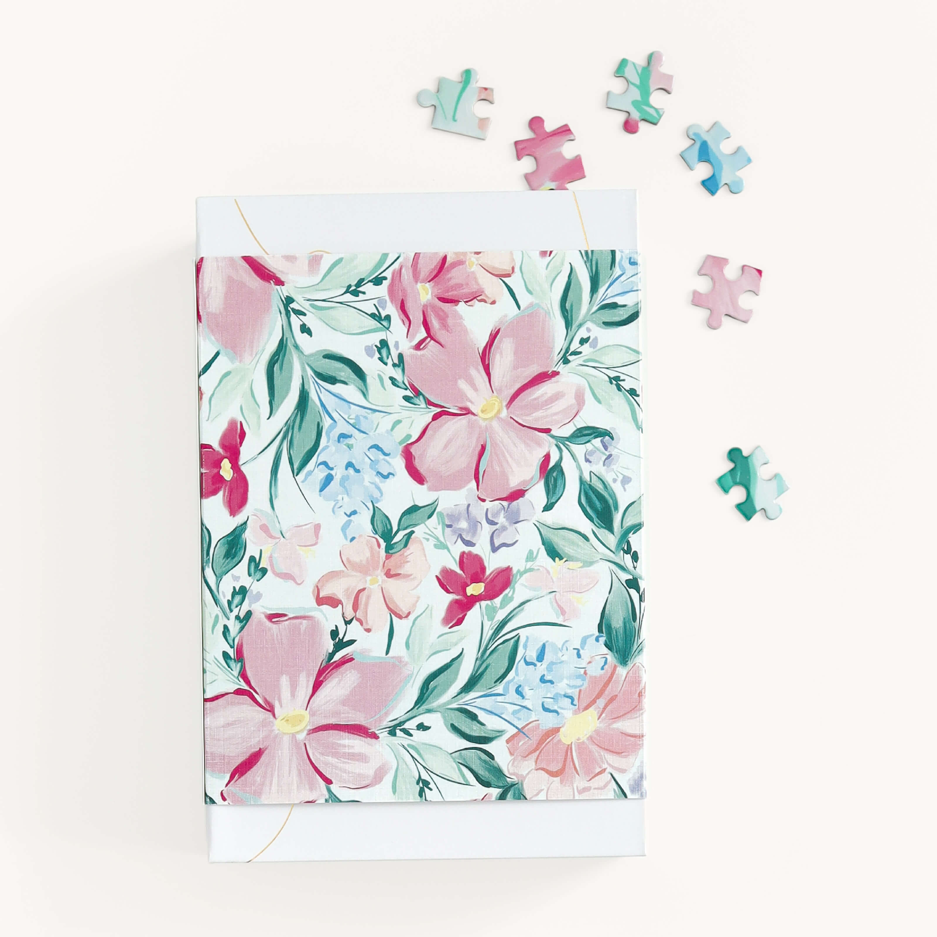 The Blush Magnolia Jiggy Puzzle by Simplified features a box adorned with watercolor flowers in pink, red, and light blue with green leaves on white. Surrounding the box are five scattered puzzle pieces that perfectly echo the floral tones in shades of pink, blue, and green.