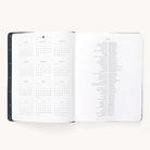 The 2024-2025 Weekly Dapperdesk Planner - Black Tie by Simplified features an open layout with a year calendar (Aug to Oct) and world holidays. Its leatherette cover with gold dots adds elegance, while high-quality paper enhances sophistication against an off-white background.