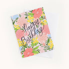 The Boxed Card Set by Simplified features a Happy Birthday card with elegant script, adorned with pink, yellow, and blue floral designs on a white background. Includes a partially visible envelope underneath.