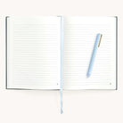 The Simplified Linen Journal - Grace is open, showing lined pages with a light blue pen resting diagonally on the right. A white ribbon bookmark peeks out from the center crease. Its chic linen hardcover sits against a plain white background, inviting creativity and thoughts.