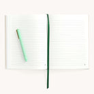 The Simplified Linen Journal - Gratitude lies open with lined pages and a linen hardcover, featuring a green ribbon bookmark in the center. A light green pen is positioned diagonally on the left page, all set against a plain white surface.