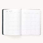 The Simplified 2024-2025 Weekly Dapperdesk Planner - Black Tie features a sleek leatherette cover and displays a monthly grid calendar for August 2024. It offers a Sunday-to-Saturday layout with blank note spaces, lined sections on the left page, and high-quality paper against a white background.