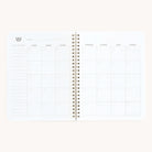 The Simplified Workbook - Finances is an open spiral-bound planner featuring a blank monthly calendar layout with Sunday to Saturday columns, perfect for tracking financial goals. A decorative emblem adorns the top left on a crisp white background.