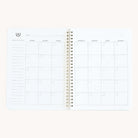 The Simplified Workbook - Project Management, an open spiral-bound planner, shows monthly spreads with daily squares. The left page includes a Focus This Month section. Weekdays from Sunday to Saturday are labeled at the top for organized planning.