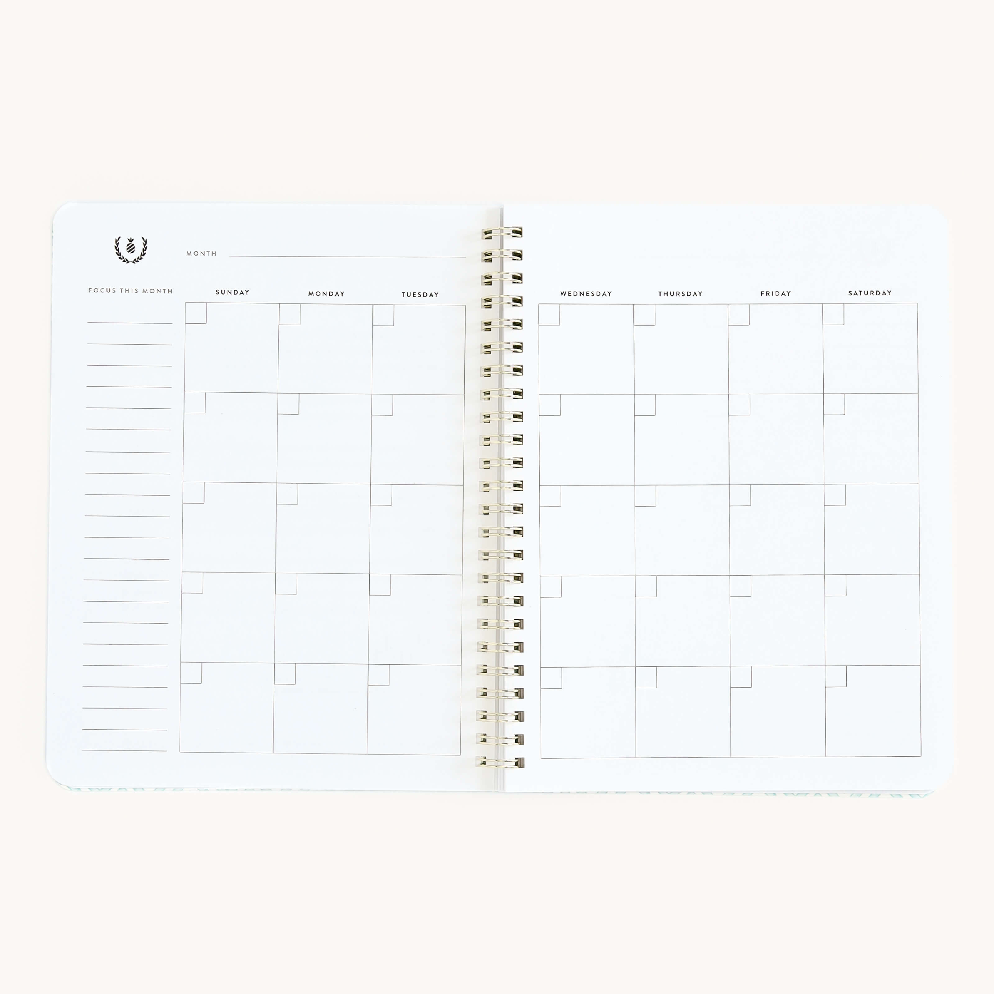 The Simplified Workbook - Project Management, an open spiral-bound planner, shows monthly spreads with daily squares. The left page includes a Focus This Month section. Weekdays from Sunday to Saturday are labeled at the top for organized planning.