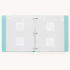 The Baby Book - Mint by Simplified is an open, light blue polka-dotted binder with blank white pages. It includes guided writing spaces, four photo placeholders, and faint decorative patterns featuring tiny heart icons in the corners.